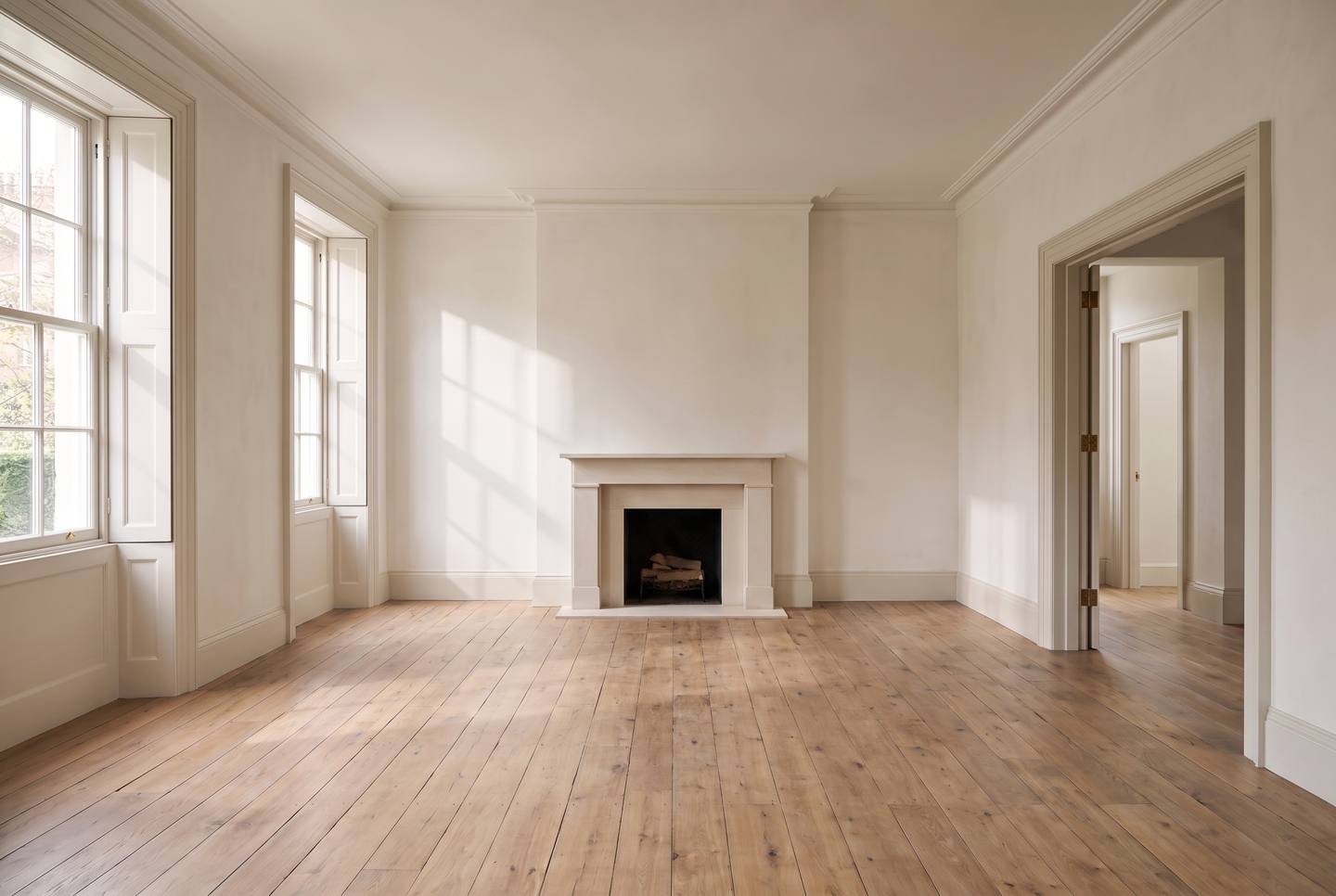 An empty Georgian living room: tall sash windows with shutters, a limestone chimneypiece, reclaimed oak boards, an open doorway on the right.