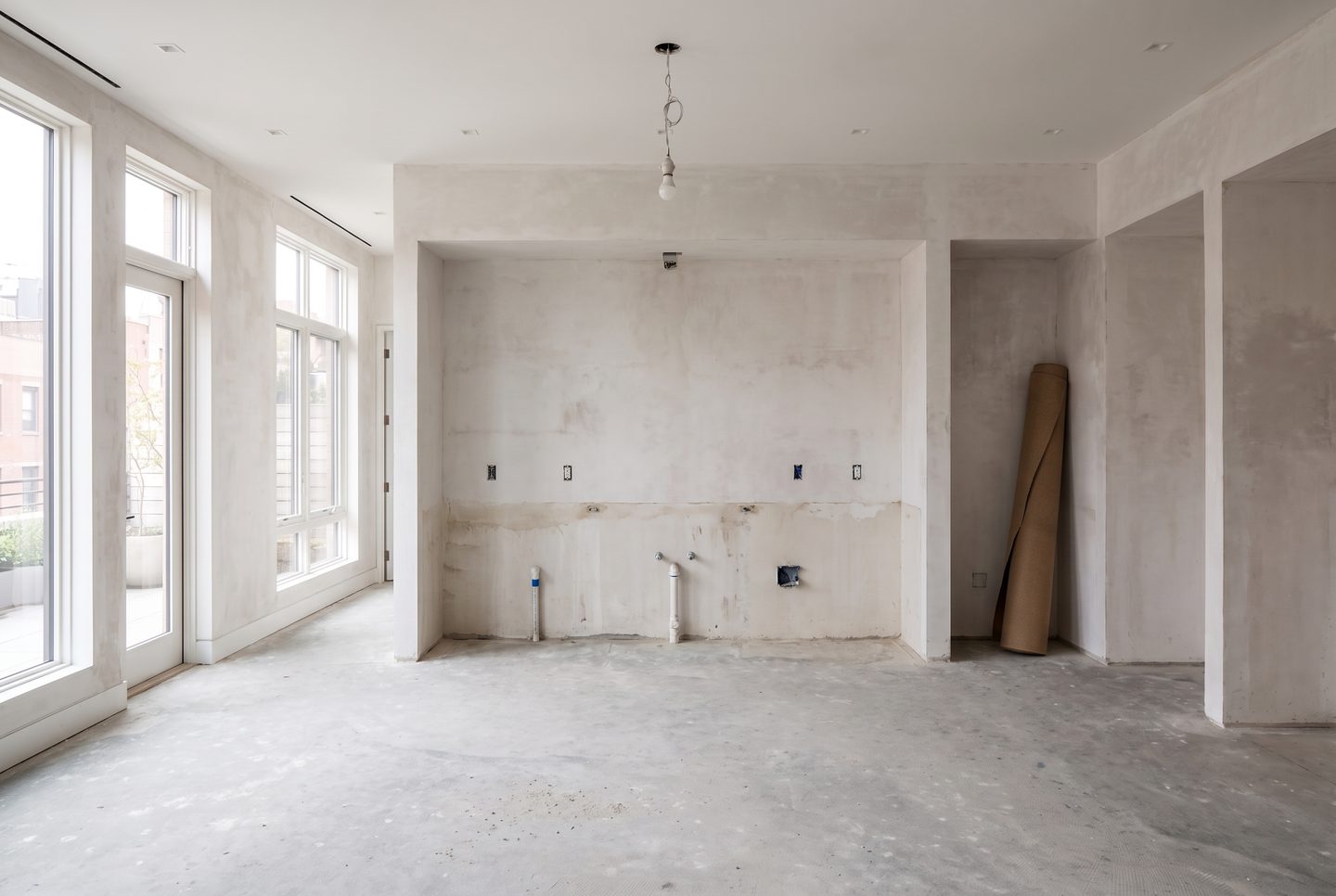 The same space as a bare shell: raw plaster, capped pipes, a concrete screed, a work bulb.